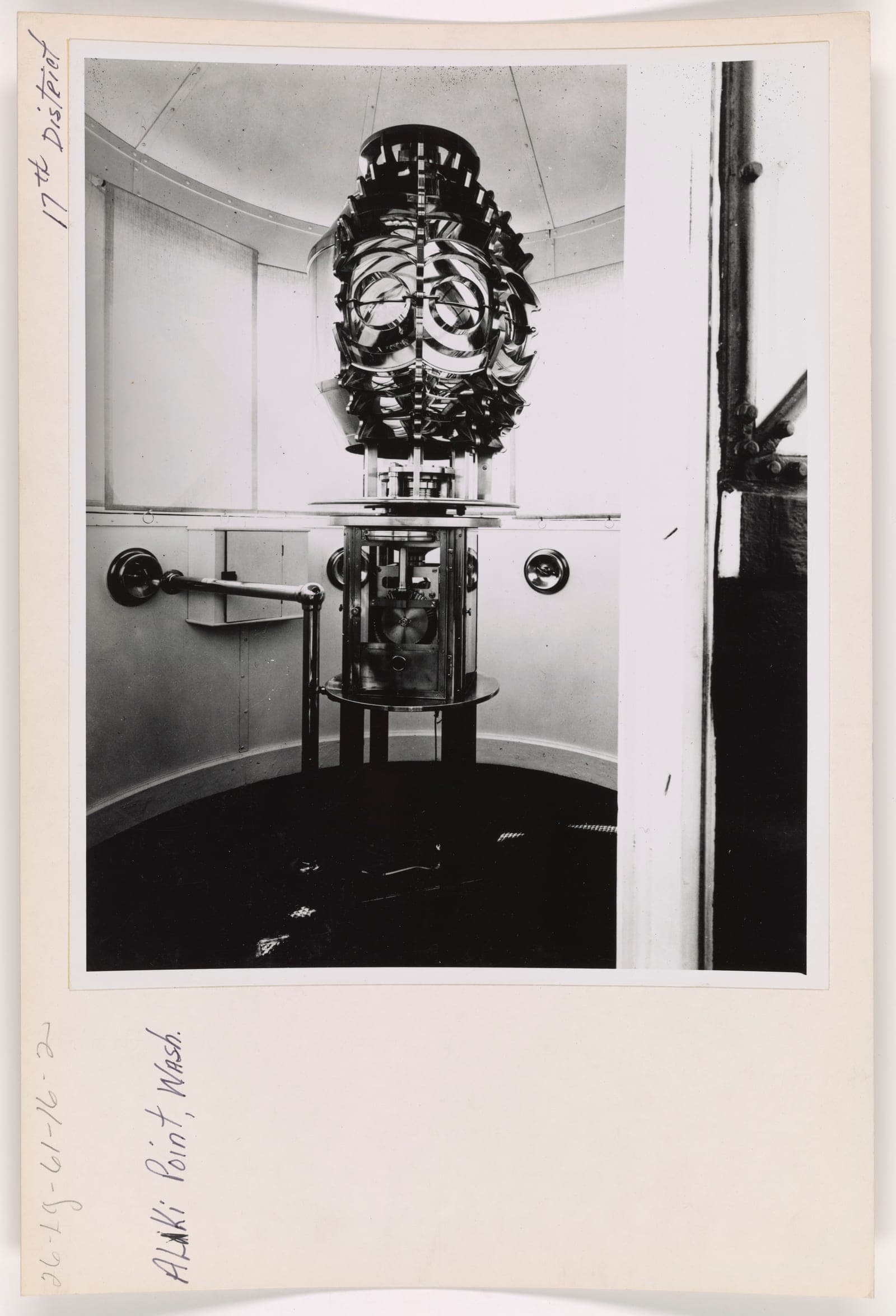 A black and white photograph of a lighthouse lantern room. A large barrel-shaped Fresnel lens made of concentric glass prisms sits on a cylindrical pedestal containing an exposed clockwork mechanism, surrounded by the curved wall and shaded windows of the lantern room.