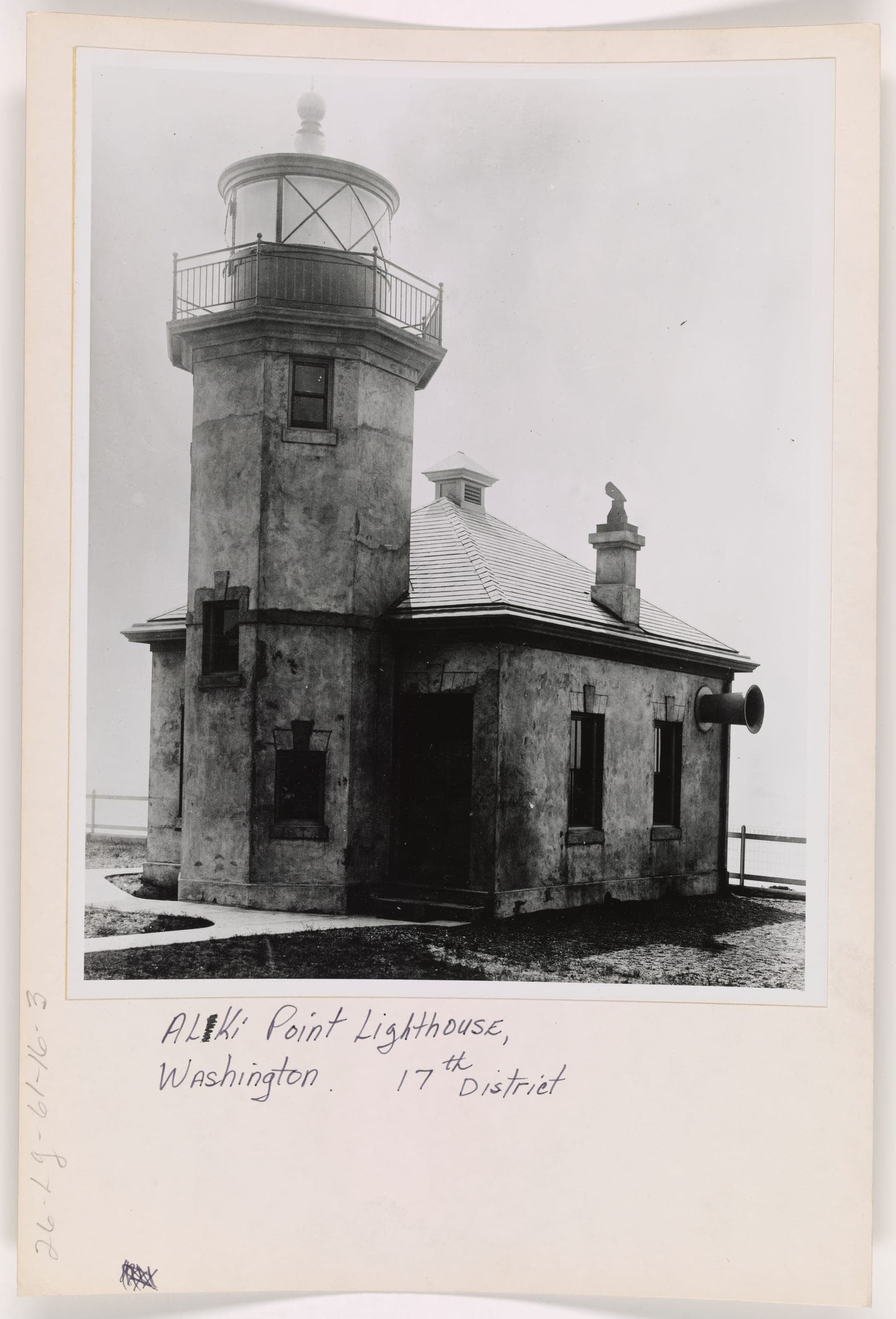 A black and white photograph, mounted on card and annotated by hand "Alki Point Lighthouse, Washington. 17th District", showing an octagonal masonry tower with a railed lantern gallery, attached to a low hipped-roof fog signal building. A trumpet-shaped fog horn projects through the wall on the right.
