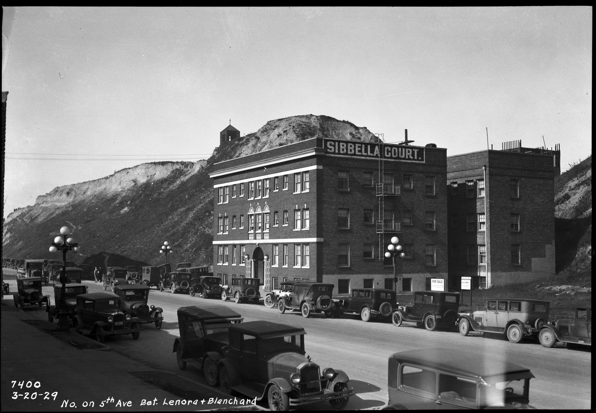 A black and white photograph of a street lined with parked 1920s automobiles. A four-story brick apartment building with "Sibbella Court" painted along its roofline, and a second brick building beside it, stand directly in front of a steep, bare earth bluff with a small building on its summit. Written on the negative: "7400, 3-20-29, No. on 5th Ave Bet. Lenora + Blanchard."